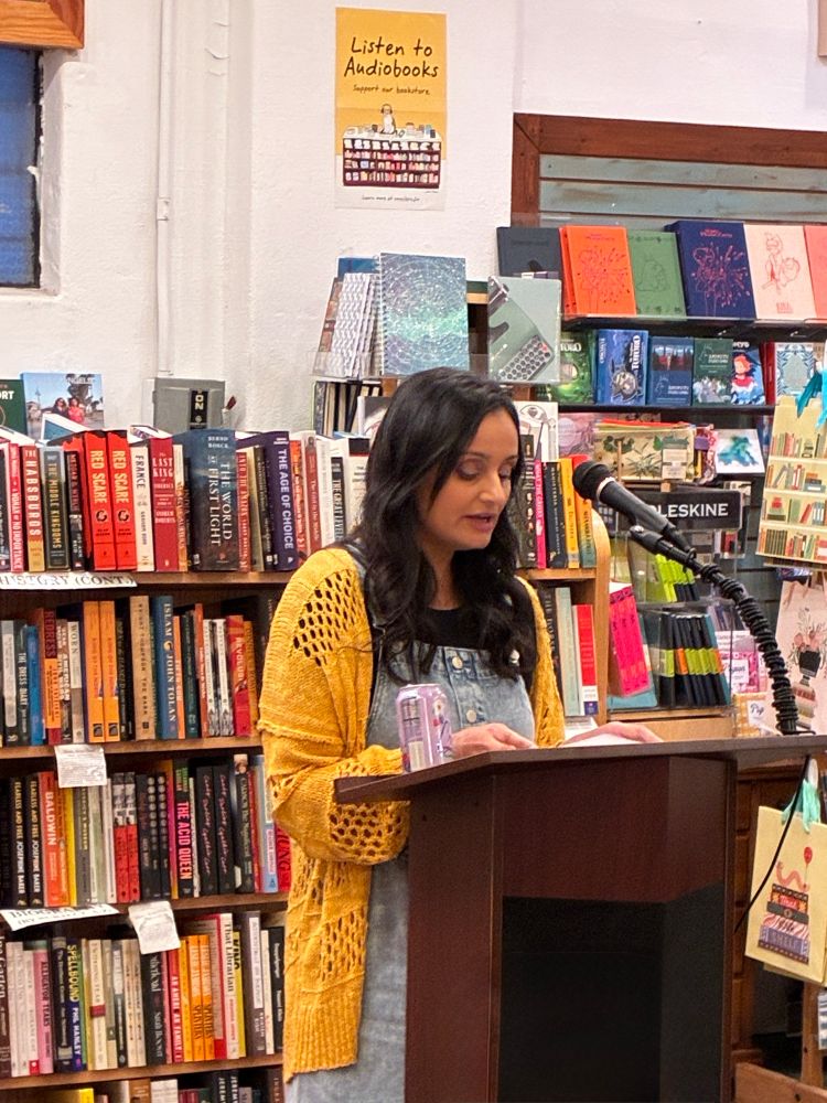 a woman in a yellow crochet cardigan and denim overalls dress over a black top standing at a podium and reading a short story for the Small Press Nite literary reading at The Book Catapult (📸 credit to @robbiemaakestad.bsky.social) 