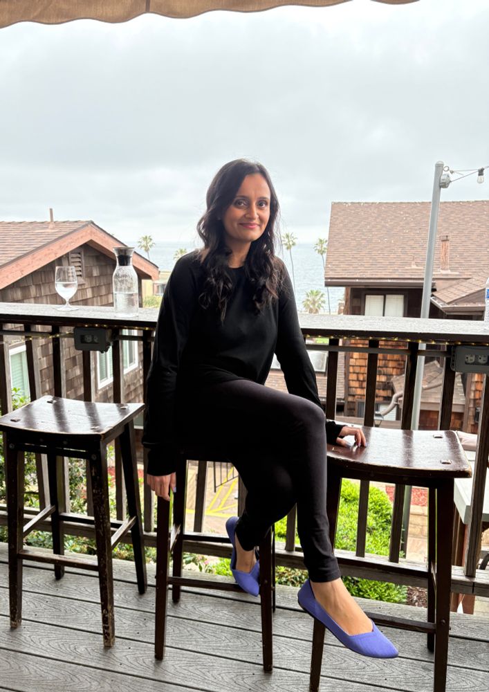 a woman—wearing a black top, black pants, and purple flats—sits on a barstool on a patio with an ocean view, having a drink before her literary reading later