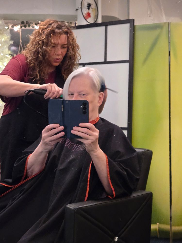 Back view of a white woman getting (from a curly-haired hairdresser) a short white, blue and black haircut meant to emulate magpies.
