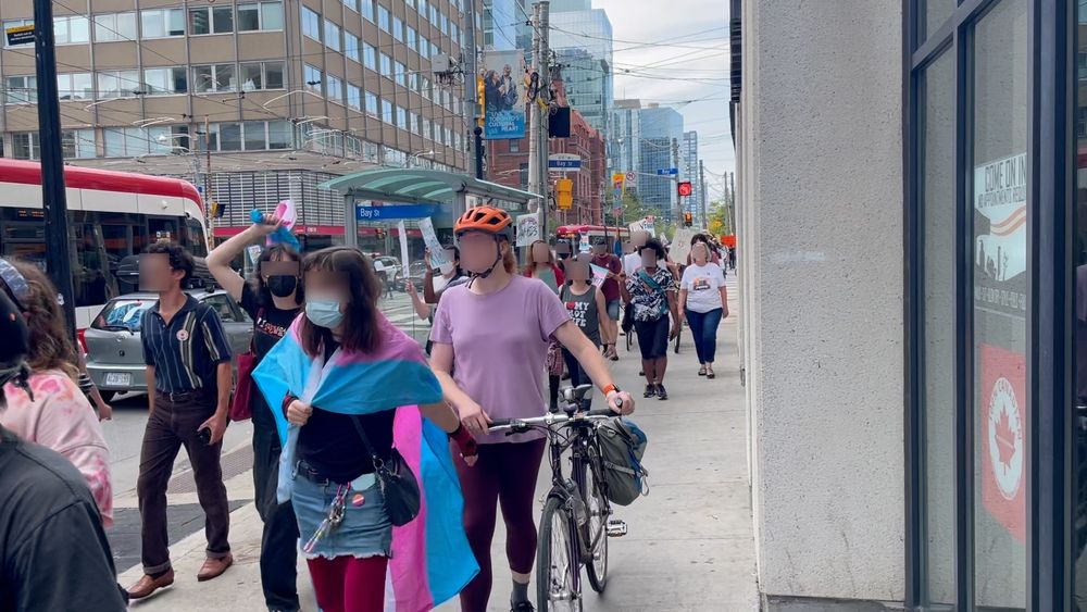 Counterprotestors march down College St east of Bay St. One demonstrator is wearing a trans flag as a cape.