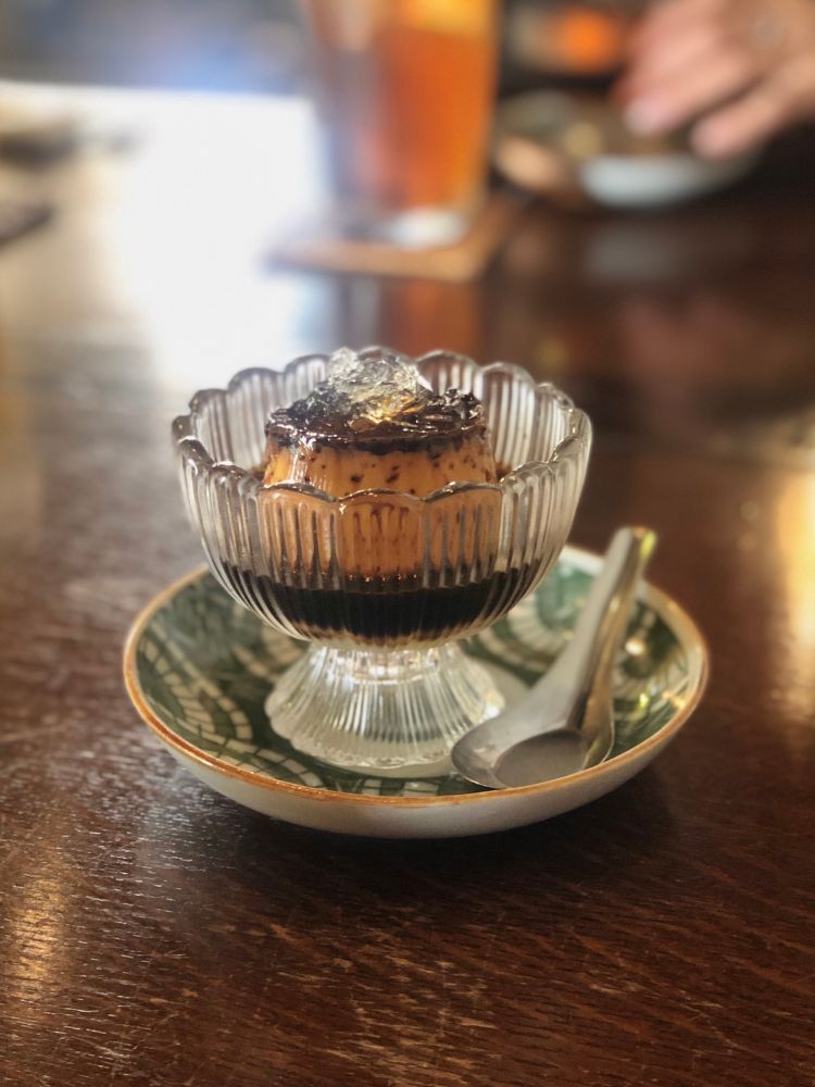 緑の柄にオレンジの縁の小皿の上にレトロなガラスの器。黄色みの強い固めプリンにコーヒーがかかっている。プリンの上には氷が乗っている