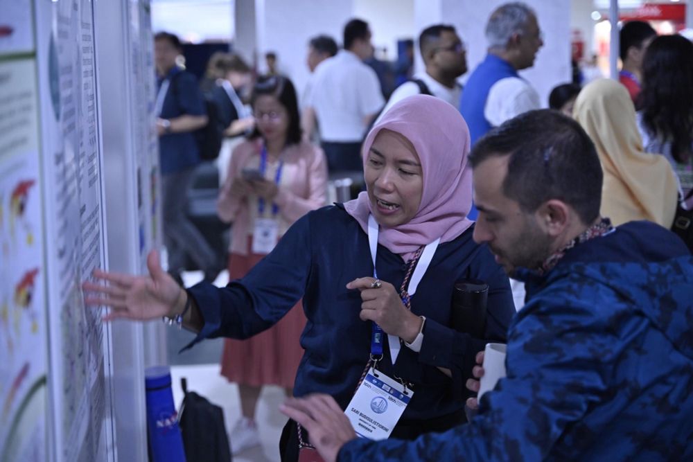 A scientist points and explains figures on a poster attached on a wall to another scientist while other scientists standing in the background. 
