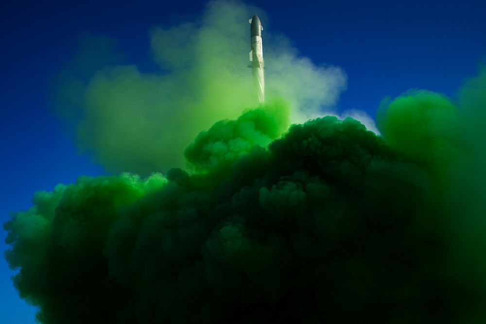 A white rocket ascends into a deep blue sky, leaving behind a thick plume of dark, toxic green smoke that billows and swirls dramatically at its base.