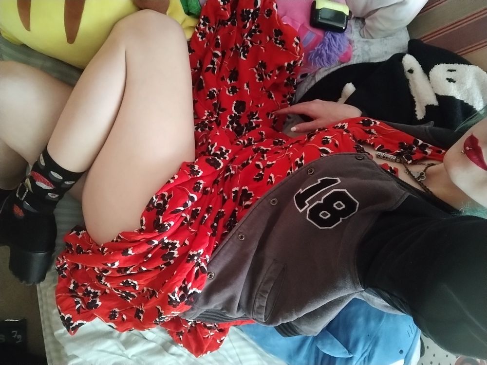 Second photo of Cricket's dress and jacket. Cricket has one knee folded, showing most of their exposed leg and their black high heel shoe. They are wearing black socks printed with sushi, and red lipstick. 