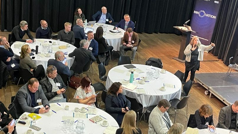 Room with round tables and people sat round it, looking at a lady speaking at the front of the room. 