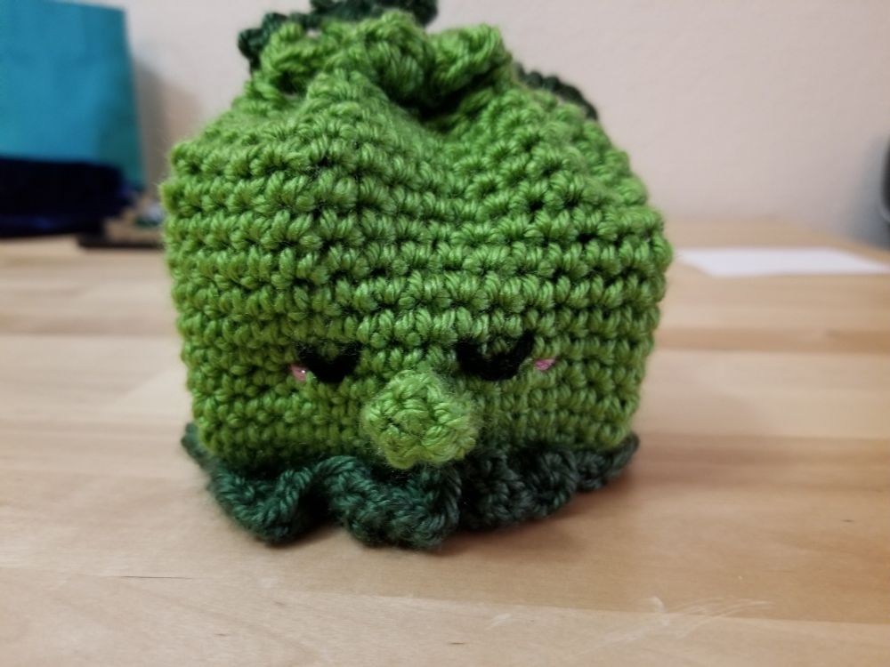 Crocheted cute green octopus bag. Has a bulbous nose and is blushing. Appears to be sleeping.