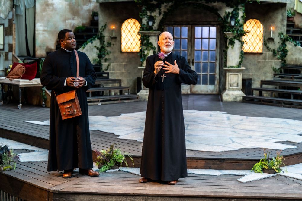 Matthew G Brown and Diego Matamoros wear black priest robes and perform a scene from Shakespeare’s Romeo and Juliet. On stage at the High Park Amphitheater in Toronto, Diego plays friar Lawrence and holds a flower, Matthew plays Friar John and  wears a brown satchel. 