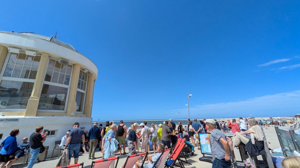 Viele Zuhörende haben sich am Borkumer Pavillon für die Lesung versammelt. Ein kleiner Durchgang wird zwischen Promande und Café gelassen