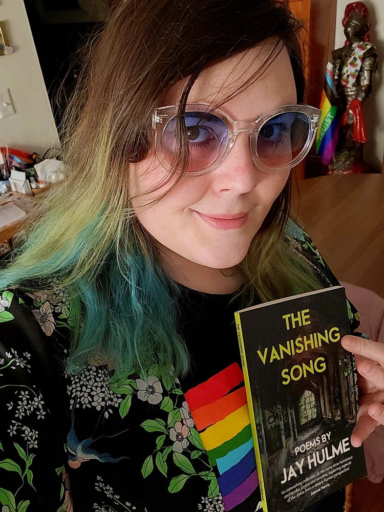 Me wearing a rainbow shirt, trans sun glasses, and a floral slip where the flowers are all trans colored. I am also holding a poetry book titled "The Vanishing zsong" by Jay Hulme.