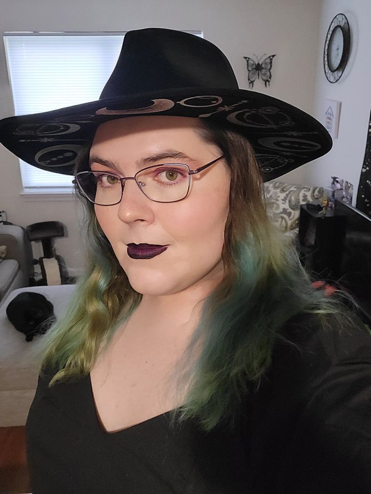 A selfie from last night. I am wearing a black dress and black hat with moon prints beneath the brim, along with black and purple lipstick. There is also a black cat in the background.