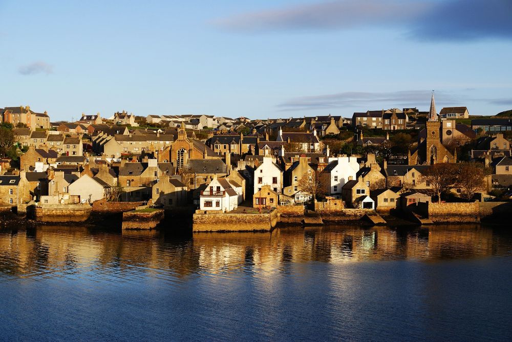 A waterfront scene of Stromness in the Orkney Islands, UK. It's a sunny day with a blue sky and streaks of high, thin clouds. Original caption from source: « The town of Stromness, Orkney Islands, Scotland, UK. The two church buildings are clearly visible. The one with the spire on the right is the former North Church converted into a Town Hall. » (📸 Geoff Wong via Wikimedia under Attribution 2.0 Generic)