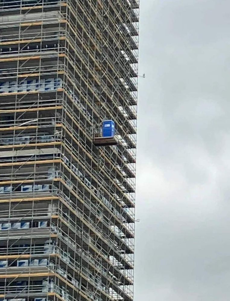long distance photo taken of the outside of a skyscraper under construction, with a small platform protruding from the exposed scaffolding supporting one blue port-a-potty over the completely empty air