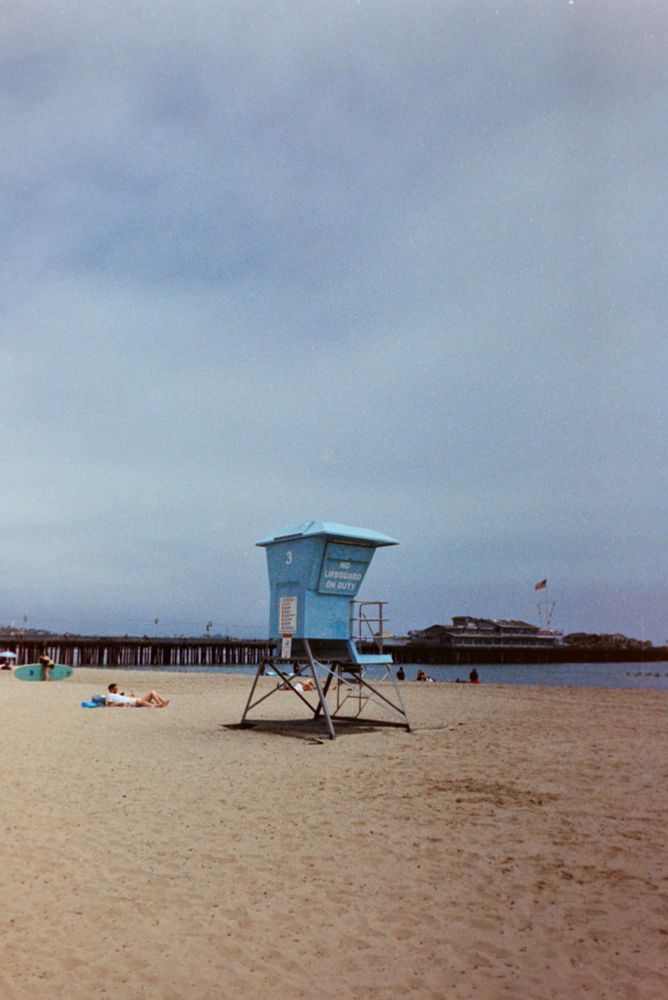 A pale blue lifeguard tower labeled “3” stands empty on a sandy beach beneath an overcast sky. The sign on the tower reads “NO LIFEGUARD ON DUTY.” A few people are lounging on the sand nearby, while a couple of paddleboards rest off to the left. In the background, a wooden pier stretches out over the ocean, with a large American flag flying above a building at the pier’s end. The scene feels quiet and mellow, capturing a calm day by the coast.