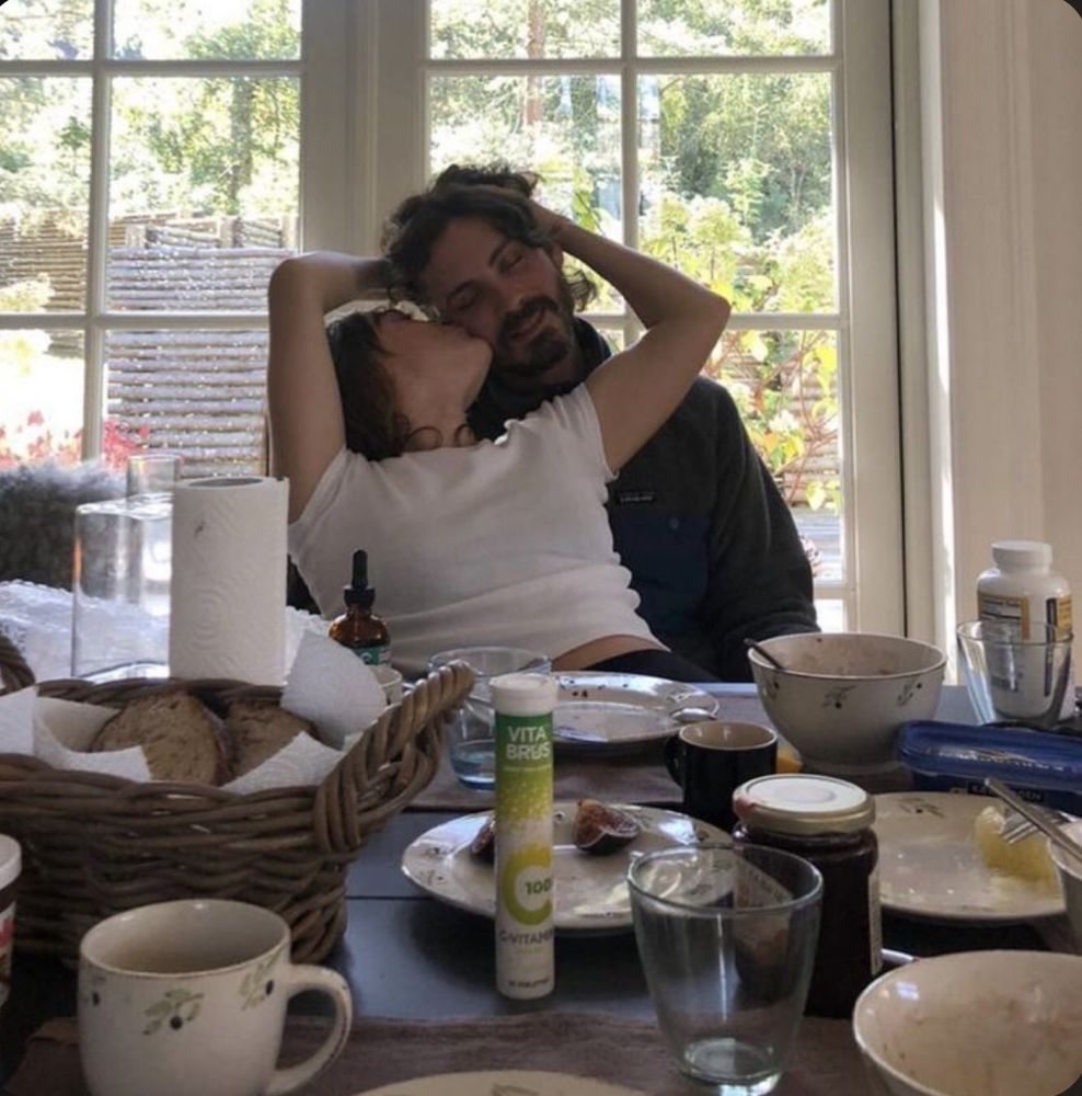 Photo of Logan Lerman and Ana Luisa Corrigan in front of a breakfast table, Ana Luisa is sat on Logan’s lap kissing his cheek and holding his head. 