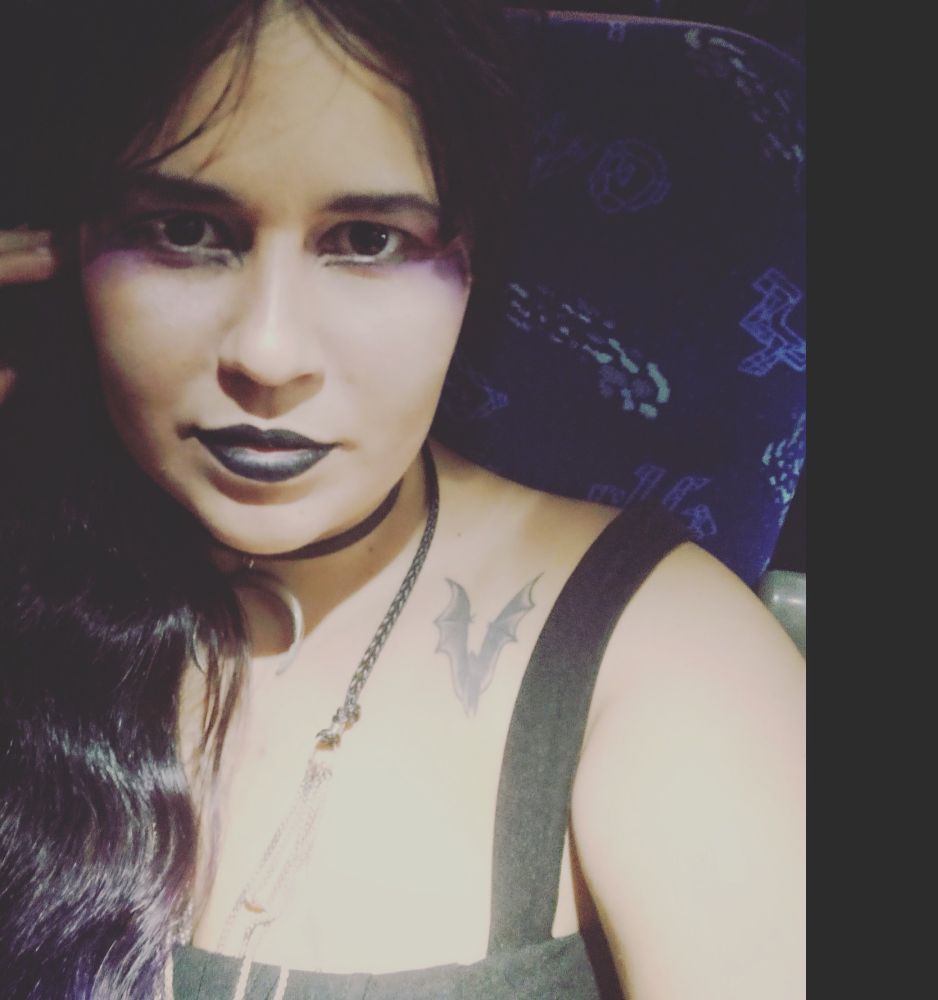 Essa sou eu. Ainda usava franja, na época. A foto está escura, com toda a iluminação na minha pele. A luz está tão forte que a tatuagem preta de morcego, que deveria destacar na pele clara, quase sumiu. Meus cabelos são castanhos, compridos, levemente ondulados e ainda dá para notar o toque de roxo que eu tinha nas pontas.