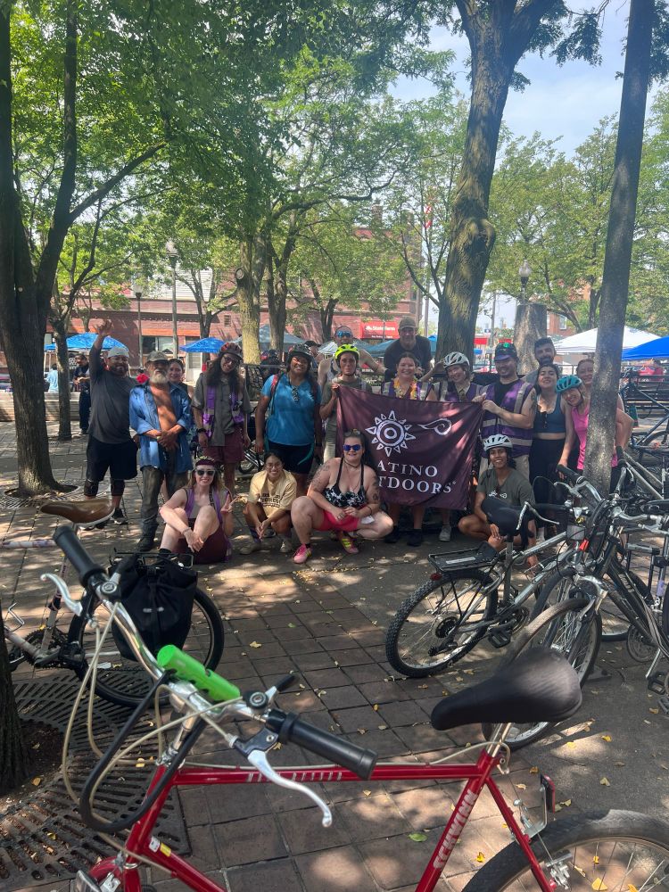 a group picture of cyclists holding up a @latinooutdoors_chicago banner under a tree canopy after a ride to Mercado de Colores❤️‍🔥 