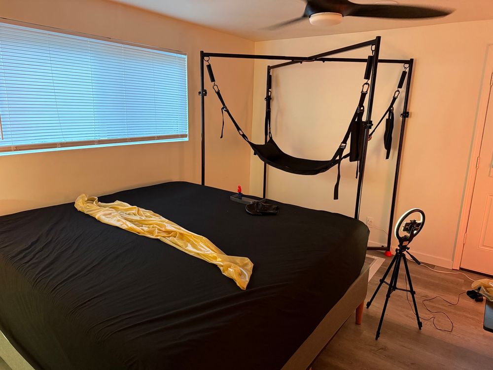 A picture of a sling next to a bed. There’s a ring light tripod set up near the foot, and a latex sleep sack on the bed. 