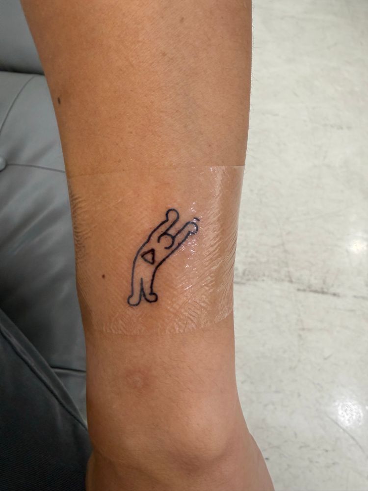 A small tattoo in the style of Keith Haring - a person with their arms raised, a triangle in their torso 