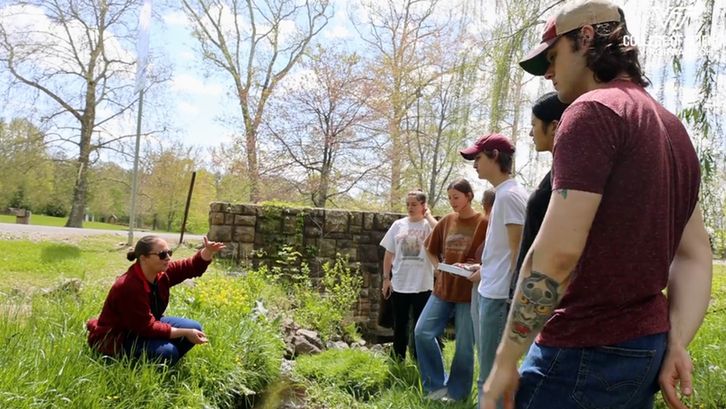 Dr. Erin Hotchkiss kneels in tall grass as she instructs undergraduate students.