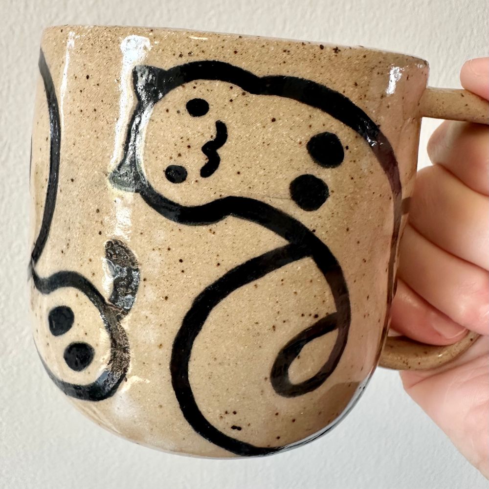 A hand (mine) holding up a handmade speckled brown stoneware mug with a “worm cat” design in black line work over bare clay with clear glaze. The cats’ long bodies loop around in fun little curlicues. 