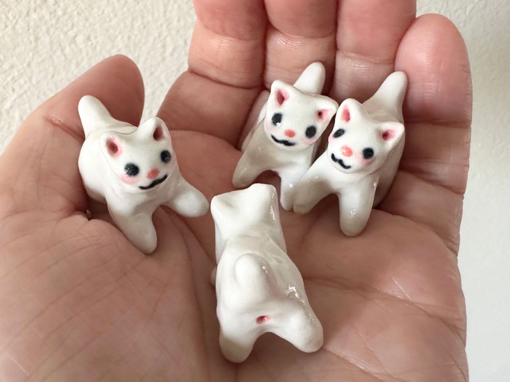 4 miniature white ceramic cats in the palm of a hand (mine). They have pink cheeks, ears, and noses. One is facing away from the camera showing they also have pink buttholes (sorry). 