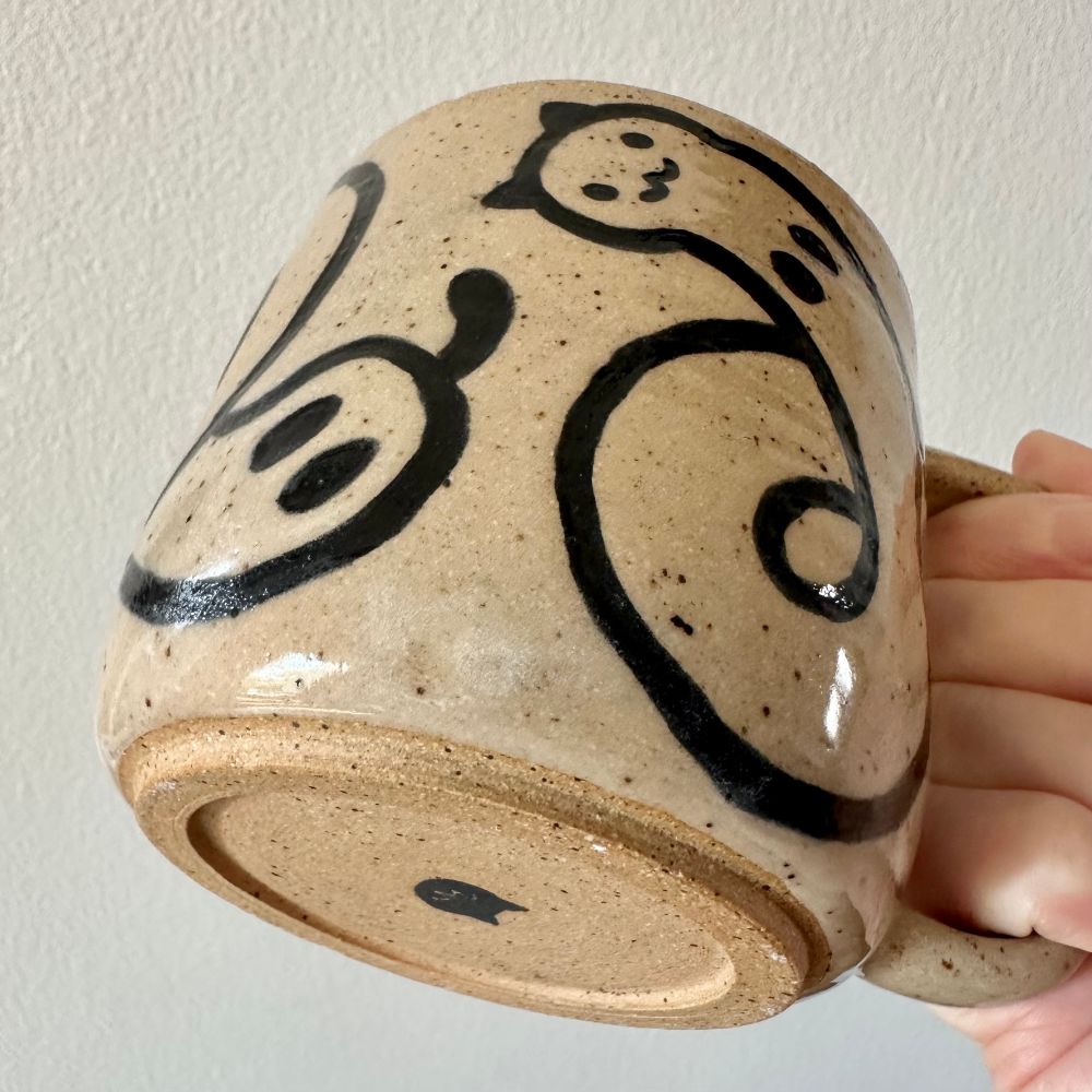 A hand (mine) holding up a handmade speckled brown stoneware mug with a “worm cat” design in black line work over bare clay with clear glaze. From this angle you can see the bottom of the mug, it has a black cat face silhouette on it. 