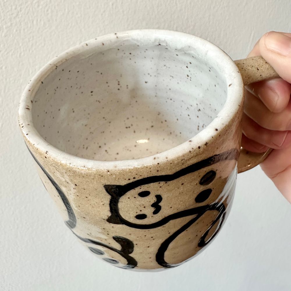 A hand (mine) holding up a handmade speckled brown stoneware mug with a “worm cat” design in black line work over bare clay with clear glaze. From this angle you can see the inside, which is white with just the brown speckles showing through. 