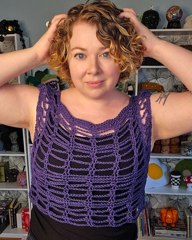 Jess wears an open weave purple crocheted top over a black outfit, standing with her hands in her hair, with shelves in the background.