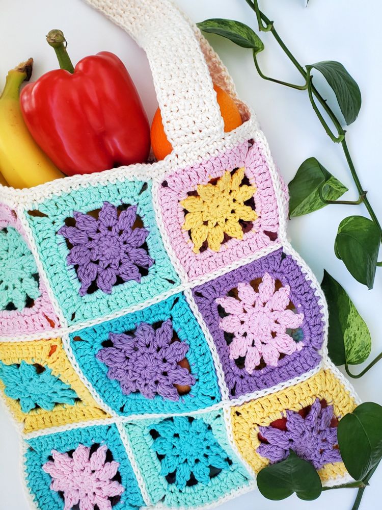 A crocheted granny square tote bag in pastel colors and filled with fruits and vegetables lays on a white background with a decorative strand of pothos leaves.