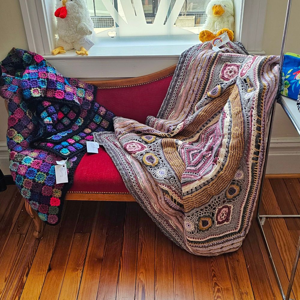 Two crocheted blankets lay on a red antique loveseat in front of a window. One blanket is made of small jewel toned granny squares with black edging. The second blanket is very detailed, including multiple styles of granny squares, in muted colors.