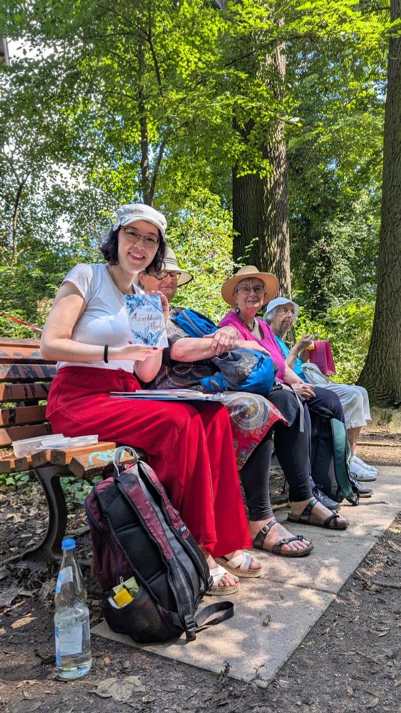 Die Autorin Jennifer Pfalzgraf halt ihr Buch "Das Azurblau deiner Worte" in den Händen. Sie sitzt mit mehreren Personen auf einer Parkbank. Die Sonne scheint und im Hintergrund sind grüne Bäume.