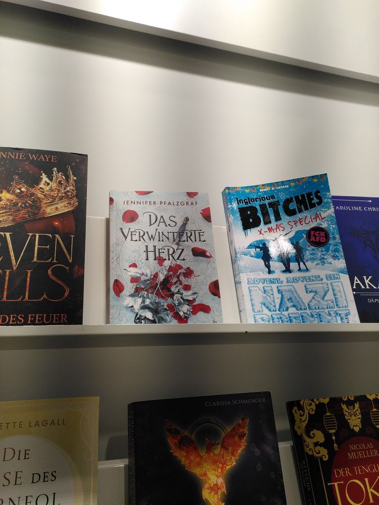 Auf einem Regal stehen mehrere Bücher nebeneinander. Zwei davon sind im Bildausschnitt abgebildet. Links das Buch "Das verwinterte Herz" von Jennifer Pfalzgraf. Rechts das Buch "Inglorious Bitches" von Benny B. Savage. 
