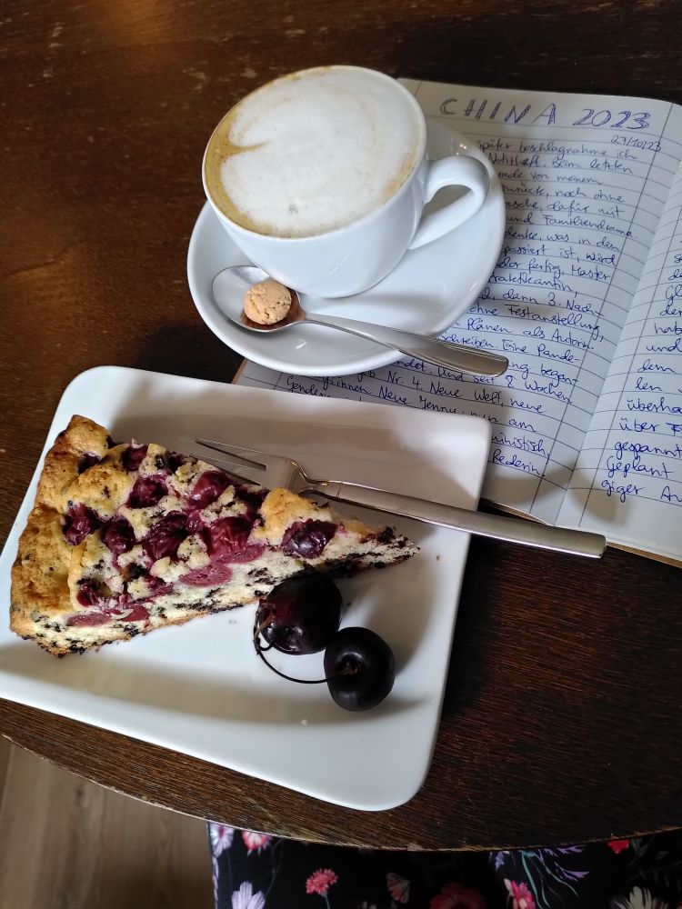 Auf einem Tisch aus dunklem Holz sind ein Teller mit Kirschkuchen, ein Cappuccino und ein aufgeschlagenes Notizbuch mit handschriftlichen Notizen zu sehen.