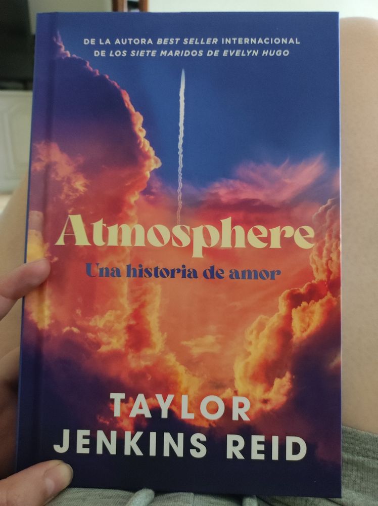Libro "Atmosphere" de la autora Taylor Jenkins Reid. En la portada salen nubes con el color anaranjado y azul del atardecer y en el medio la estela de un cohete despegando. 