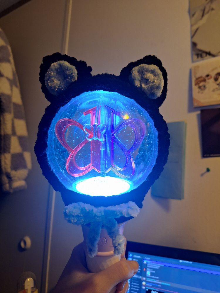 picture of a VIVIZ lightstick with a crochet cover that is black with cat ears that are light blue in the center. it also has a light blue bow in the center.