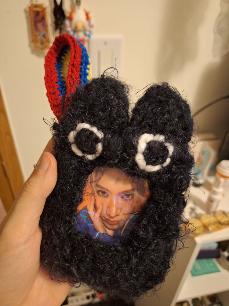 A fuzzy crocheted photocard holder resembling a black cat with wide eyes and a multicolored strap extending from the top left corner. There is a photocard of the idol Ten from Wayv inside.