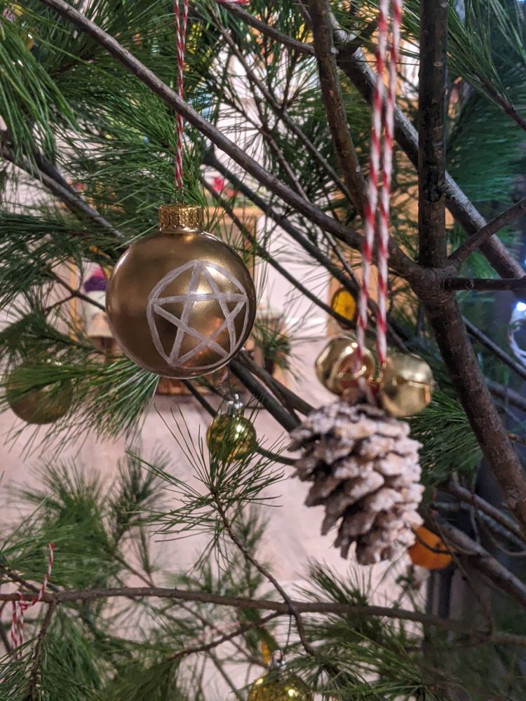 A few different ornaments on a pine tree, including a bauble with a pentacle, jingle bells, and frosty pinecones 