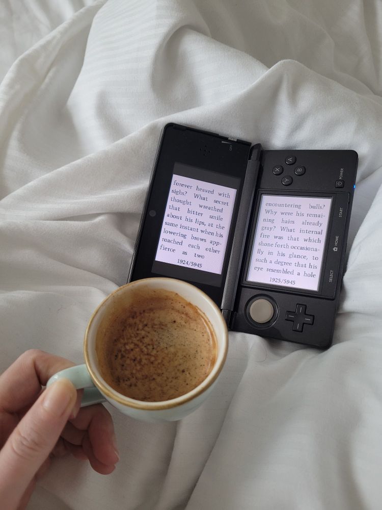 A hand holds a small coffee and a Nintendo 3DS rests on a white bedsheet, displaying text from The Hunchback of Notre Dame.