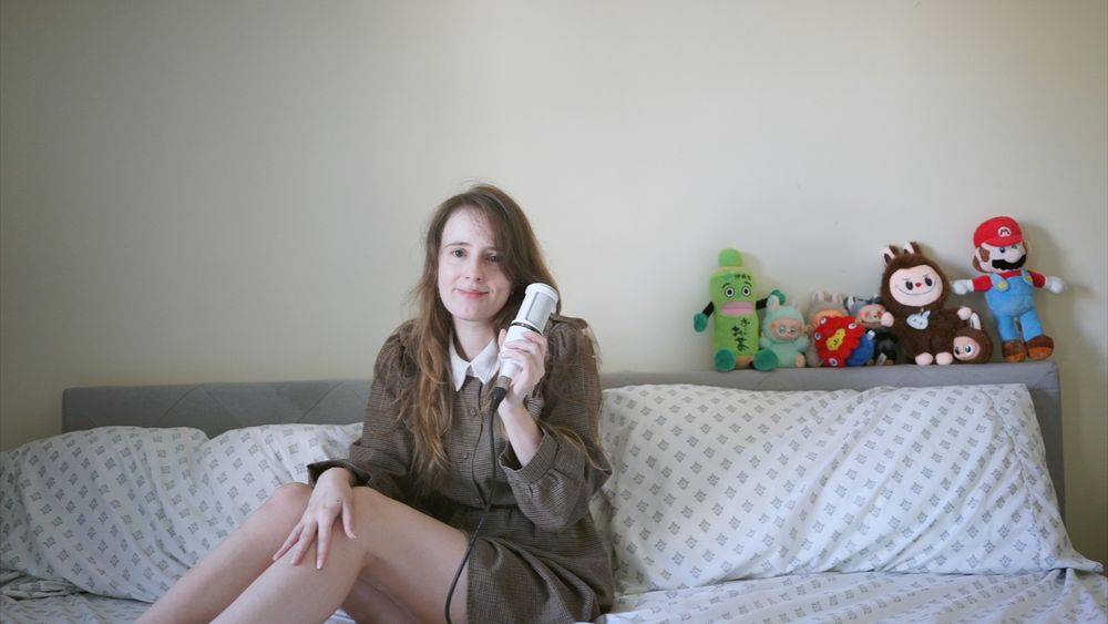 A woman smiles serenely, sitting on a bed and holding a microphone next to several labubu type items.