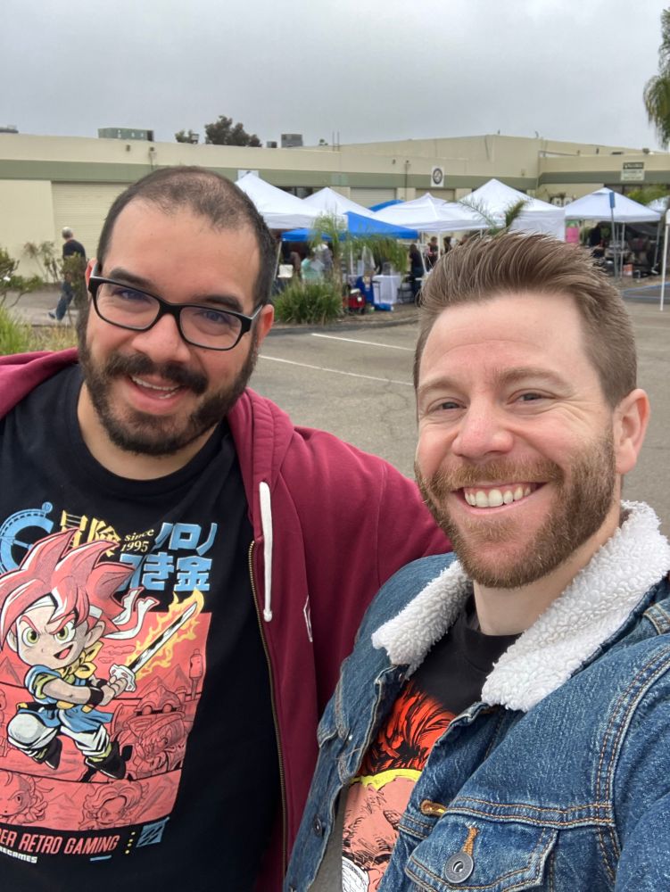 Two smiling men pose for a selfie outdoors in front of a small pop-up market or event with white tents and palm trees. One wears a red hoodie and Chrono Trigger-themed shirt, and the other wears a denim jacket with a fur collar and a Beta Ray Bill and Juggernaut graphic tee. The sky is overcast.