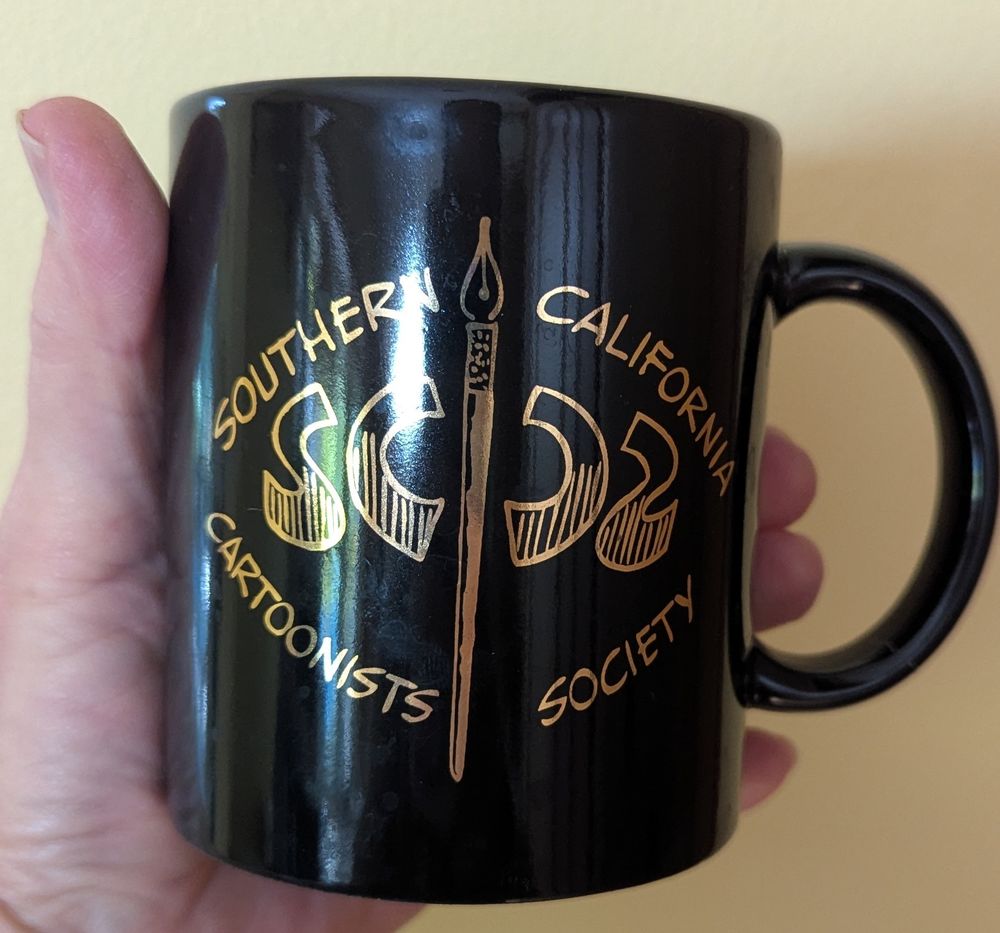 Close-up photo of a hand holding a black ceramic mug inscribed with gold lettering: Southern California Cartoonists Society /SCCS. Photo by Ellen Friedman