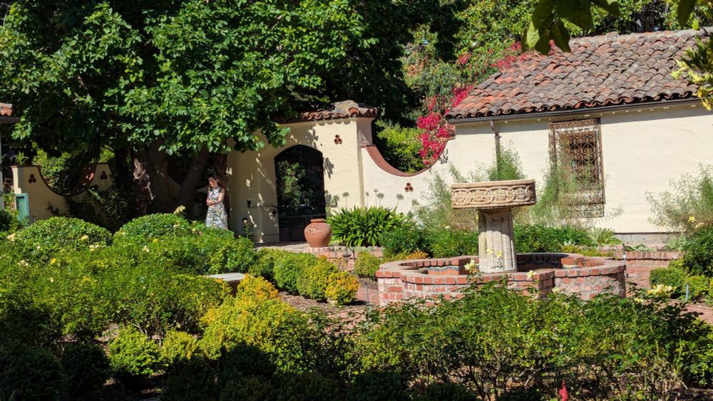 Courtyard of Allied Arts Guild in Menlo Park, CA seen in bright midday sun. Low, trimmed green shrubs surround a brick walkway and fountain. Behind it is a light stucco wall with tile roof and bright red bougainvillea. Photo by Ellen Friedman 30 Aug 2025. 