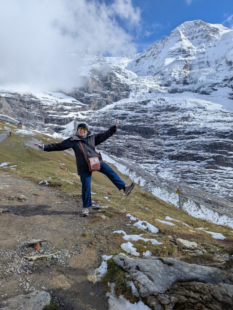 Woman hiker being silly on a mountain trail, standing on one leg with arms outstretched and big smile. Slope at edge of trail is green and patches of rock and snow. Behind, in near background, is looming snow-covered peak called the Monch.  Photo by Ellen Friedman. 30 Sept 2025