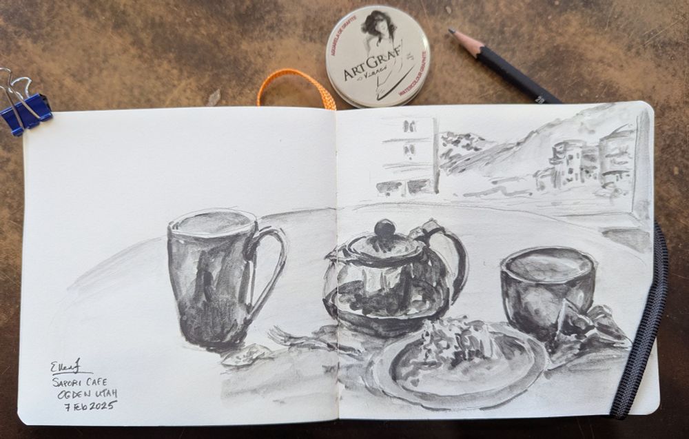 Black and white drawing and small sketchbook of a cafe table with teacup teapot and a coffee mug, distant mountains in the background beyond the town. Drawing by Ellen Friedman