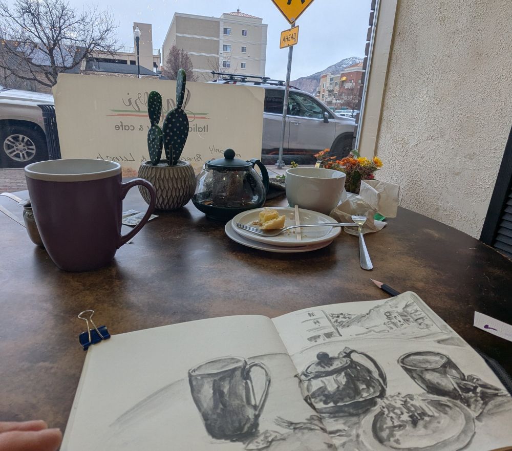 Cafe table with teacup teapot and a coffee mug, distant mountains in the background beyond the town. Sketchbook in foreground. Photo by Ellen Friedman