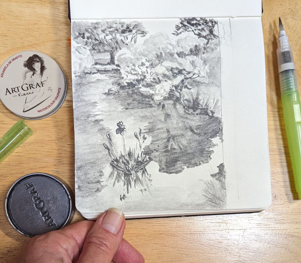 On left of photo, ArtGraf soluble graphite: round cake in a tin that is open, with lid showing name above. A small sketchbook sketch of pond in Japanese garden is held by artist's hand. On right is a water brush pen used to make this small painting. Photo and painting by Ellen Friedman