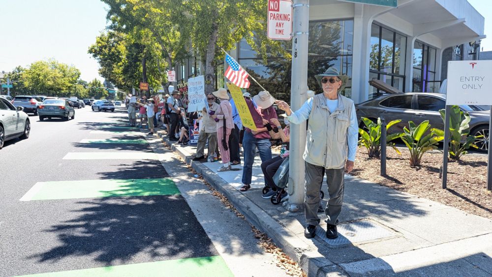 Pro-democracy anti-trump protesters line a busy street, waving American flags and holding signs.