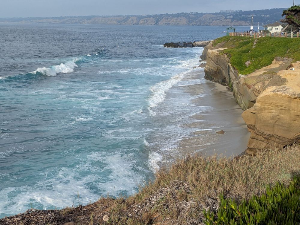 View of Pacific Ocean with beautiful turquoise colored water and white caps. Crashing onto a sand beach below. Sandstone cliffs. Upper right some dark rocks jut out into the water. Photo by Ellen Friedman Nov 2025