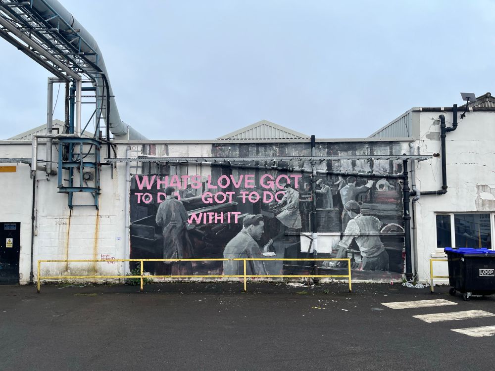 Graffiti mural saying “what’s love got to do with it?” With a depiction of the paper factory in the past in the background 
