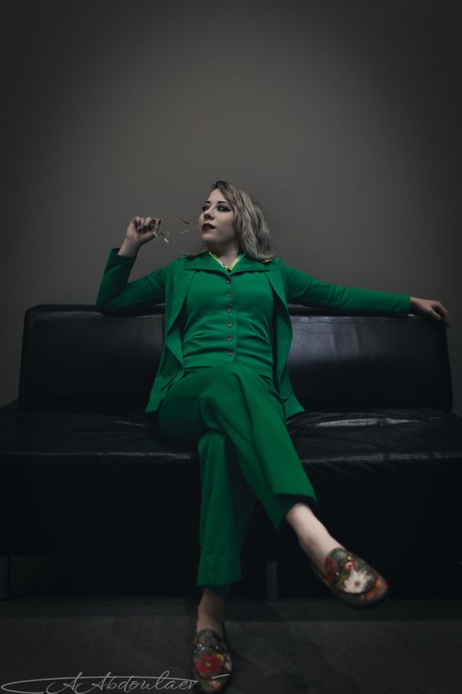 Woman in a green suit seated on a leather sofa, plain gray background. Desaturated. Even lighting.